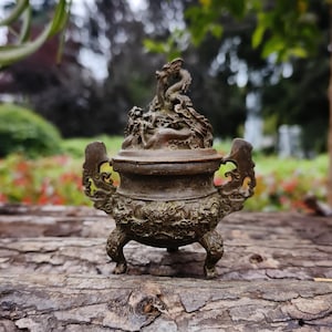 May include: A bronze incense burner with intricate dragon and floral relief details. The burner has a lid with a dragon sculpture on top, two handles, and three small feet. The overall design is ornate and detailed.