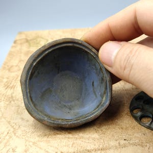 May include: A close-up of a small, antique metal container with a rounded shape. The interior is a muted blue, contrasting with the darker, aged exterior. The container is held by a hand, with a separate, circular piece visible.