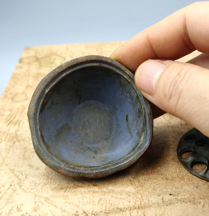 May include: A close-up of a small, antique metal container with a rounded shape. The interior is a muted blue, contrasting with the darker, aged exterior. The container is held by a hand, with a separate, circular piece visible.