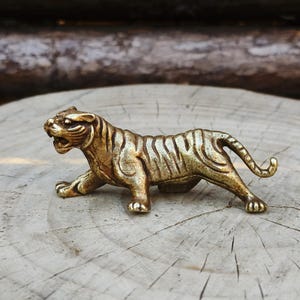 Puede incluir: Pequeña y detallada figura de tigre de latón en posición agachada. El tigre tiene rayas visibles y está sobre un fondo de madera natural. La figura mide aproximadamente 7.6 cm de largo.