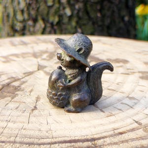 May include: A bronze squirrel figurine wearing a hat and holding an acorn. The squirrel has a detailed texture and is posed on a wooden surface. The figurine is a decorative item, possibly for a home or garden.