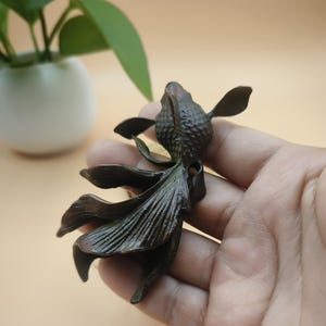 May include: A bronze-colored sculpture of a fish with flowing fins. The fish is held in a hand, with a blurred background featuring a white vase and green plant. The sculpture has detailed scales and fins, and is likely a decorative item.