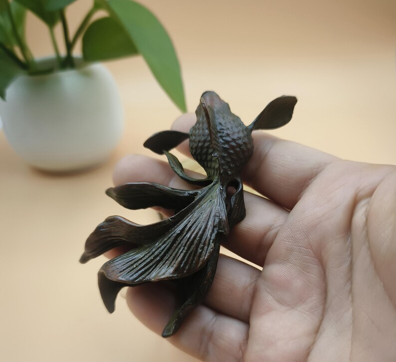 May include: A bronze-colored sculpture of a fish with flowing fins. The fish is held in a hand, with a blurred background featuring a white vase and green plant. The sculpture has detailed scales and fins, and is likely a decorative item.