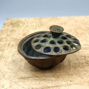 May include: A bronze-coloured lotus flower-shaped incense burner with a perforated lid. The lid is a light green colour and has a small, round handle. The incense burner is sitting on a light brown surface.
