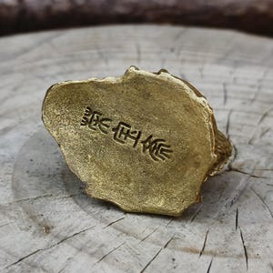 May include: A small, gold-colored figurine with a textured surface. The base of the figurine has embossed Japanese characters. The figurine is resting on a wooden surface.