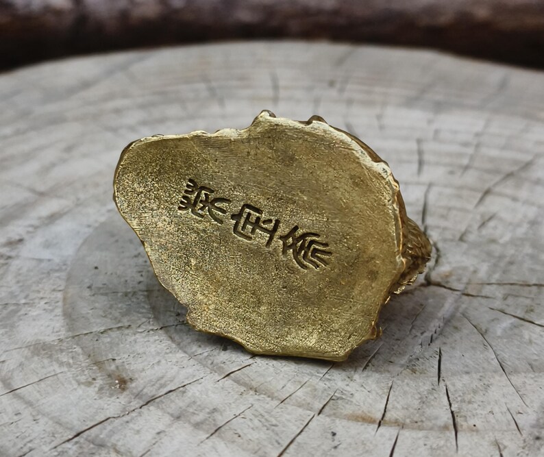 May include: A small, gold-colored figurine with a textured surface. The base of the figurine has embossed Japanese characters. The figurine is resting on a wooden surface.