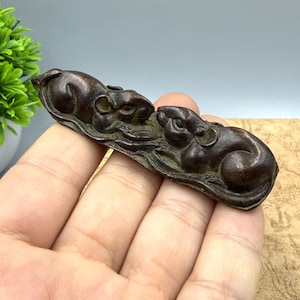 May include: A bronze-colored sculpture featuring two stylized rats resting on a textured base. The rats are depicted in a relaxed pose, with detailed features. The sculpture is held in a hand, suggesting its size and portability. The overall aesthetic is antique.