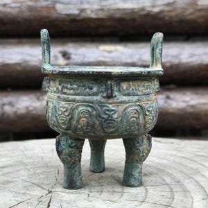 May include: An antique bronze vessel with a weathered patina. The vessel features intricate relief carvings and two handles. It stands on three legs, each with a detailed design. The color is a mix of green and brown, typical of aged bronze.