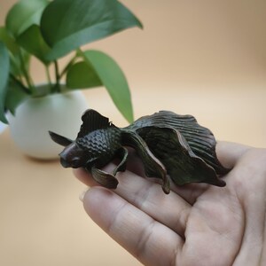 May include: A bronze-colored goldfish sculpture with detailed fins and tail. The sculpture is held in a hand, showcasing the intricate design and texture of the fish. The background includes a potted plant with green leaves.