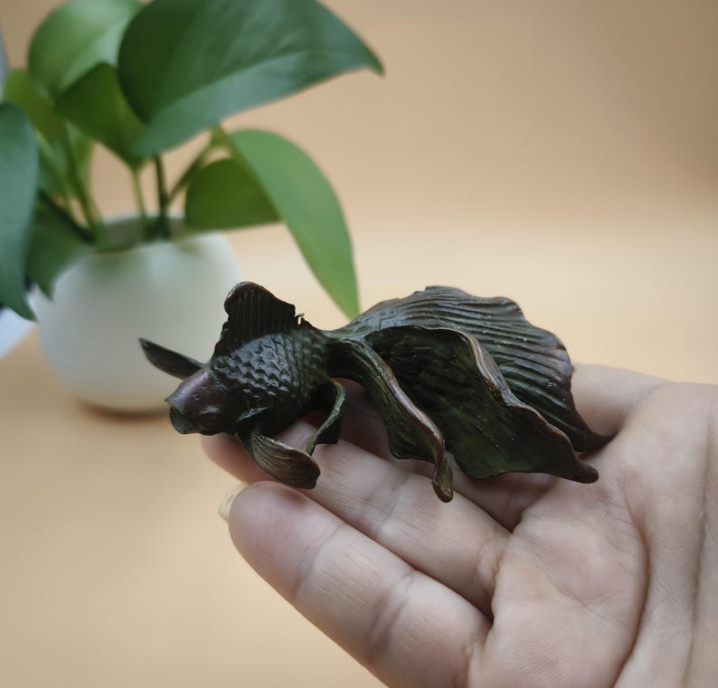 May include: A bronze-colored goldfish sculpture with detailed fins and tail. The sculpture is held in a hand, showcasing the intricate design and texture of the fish. The background includes a potted plant with green leaves.