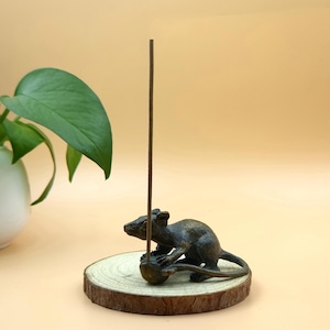 May include: A bronze rat incense holder on a wooden base. The rat is sculpted in a realistic pose, holding a small ball. A brown incense stick is inserted into the ball. The base is a round slice of wood with the bark still attached.