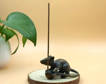 May include: A bronze-colored rat incense holder with a wooden base. The rat is sculpted in a realistic pose, holding a small ball. A brown incense stick is inserted into the ball. The base is a round slice of wood with the bark still attached.
