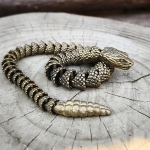 May include: An articulated brass snake figurine. The snake is coiled, with a textured body and a detailed head. The brass has a warm, antique finish. The snake is resting on a weathered, light-coloured wooden surface.