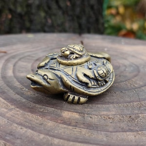 Könnte beinhalten: Eine detaillierte Bronze-Schildkrötenfigur mit einer kleineren Schildkröte auf dem Rücken und einer weiteren an der Seite. Die Figur hat detaillierte Schalen- und Hautmerkmale und steht auf einer Holzoberfläche. Das Design ist eine detaillierte Skulptur im antiken Stil.