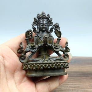 May include: A detailed bronze statue of a deity with multiple arms and ornate headdress. The figure is in a seated position with hands clasped in prayer. The statue has intricate detailing and a dark, aged patina, suggesting its age. The base features a decorative floral pattern.