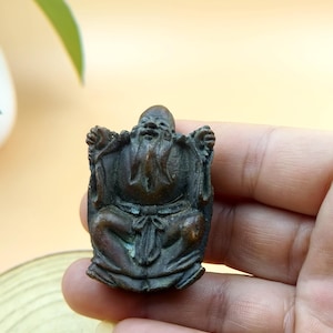 Puede incluir: Pequeña figura de color bronce de un anciano sentado y sonriente, con una larga barba y vestiduras tradicionales. La figura tiene rasgos faciales detallados y se sostiene en una mano.