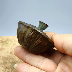 May include: A bronze-colored, decorative sculpture of a lotus seed pod. The pod has a textured surface with vertical lines and a small, detailed stem. The sculpture is held in a hand, showcasing its size and form.