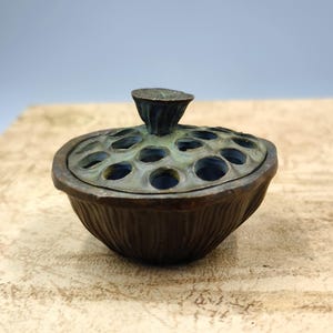 May include: A bronze-colored incense burner with a lotus pod design. The lid features a series of circular openings and a small, stylized lotus flower handle. The base has a ribbed texture, and the overall appearance is antique.