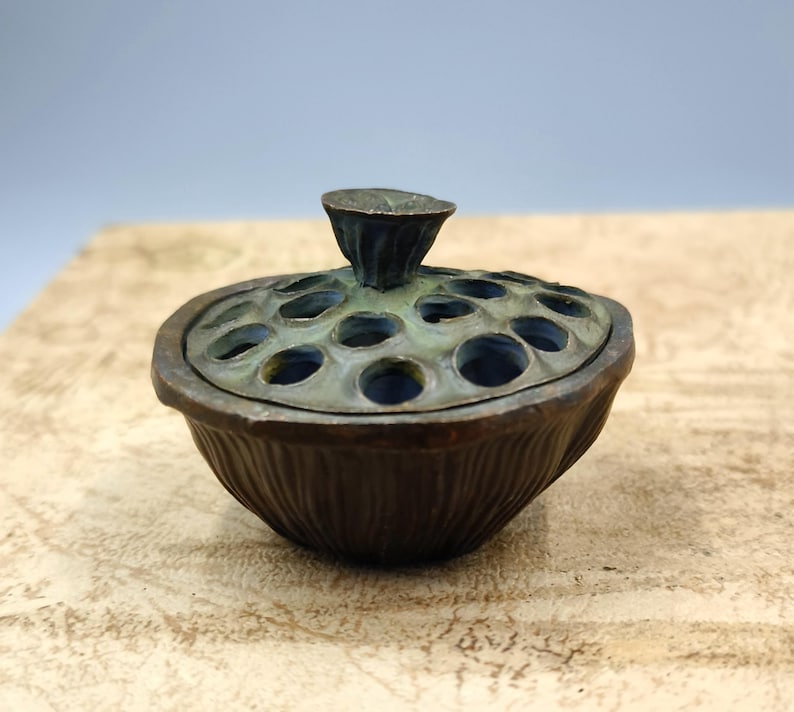May include: A bronze-colored incense burner with a lotus pod design. The lid features a series of circular openings and a small, stylized lotus flower handle. The base has a ribbed texture, and the overall appearance is antique.