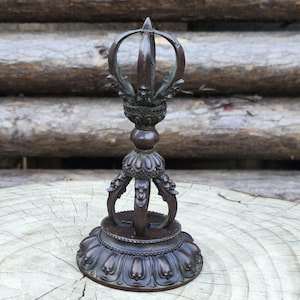 May include: A bronze-coloured, ornate Vajra, a ritual object with a central pronged sphere, a bulbous midsection, and a lotus-shaped base. The top features a crown-like design. The object is placed on a wooden surface.