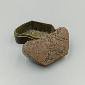 Op de afbeelding: Een open antieke doos met een koperkleurig deksel. Het deksel heeft een gegraveerde afbeelding van een zittende figuur in een landschap. De doos heeft een donkerbruine basis en een goudkleurig interieur.