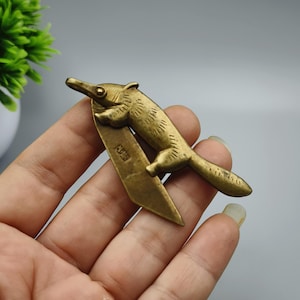 Puede incluir: Un marcapáginas de metal color bronce con un diseño de oso hormiguero. El oso hormiguero está representado aferrado a un trozo de metal rectangular. El marcapáginas tiene una estética vintage y se sostiene en una mano.