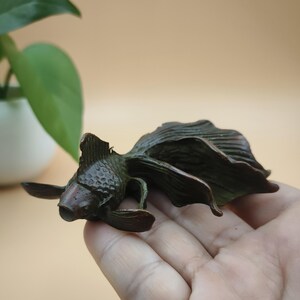 May include: A bronze-colored goldfish sculpture with detailed fins and scales. The sculpture is lying on its side, showcasing its flowing tail and delicate features. The artwork is a decorative piece, likely for home decor.