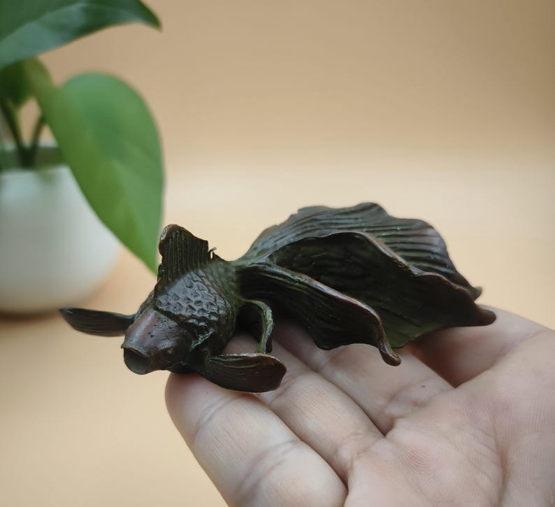 May include: A bronze-colored goldfish sculpture with detailed fins and scales. The sculpture is lying on its side, showcasing its flowing tail and delicate features. The artwork is a decorative piece, likely for home decor.