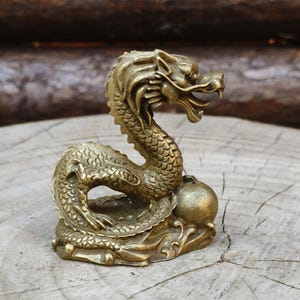 May include: A detailed brass dragon figurine with an open mouth and intricate scale details. The dragon is coiled and rests on a base with a sphere. The figurine is a golden color and is set against a natural wood background.