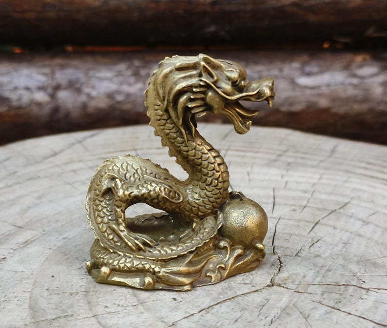 May include: A detailed brass dragon figurine with an open mouth and intricate scale details. The dragon is coiled and rests on a base with a sphere. The figurine is a golden color and is set against a natural wood background.