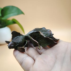 May include: A bronze-colored goldfish sculpture with detailed fins and tail. The sculpture is held in a hand, showcasing the intricate design and texture of the fish. The sculpture is a decorative item.
