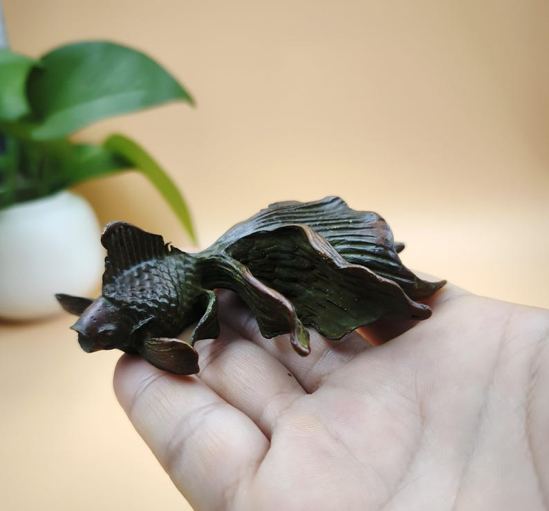 May include: A bronze-colored goldfish sculpture with detailed fins and tail. The sculpture is held in a hand, showcasing the intricate design and texture of the fish. The sculpture is a decorative item.