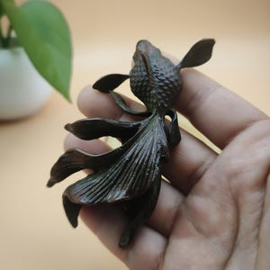 May include: A bronze-colored sculpture of a goldfish, held in a hand. The fish has detailed fins and scales, with a textured body. The sculpture is a decorative item, likely for home decor or as a collectible.