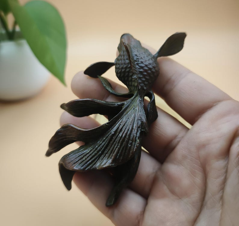 May include: A bronze-colored sculpture of a goldfish, held in a hand. The fish has detailed fins and scales, with a textured body. The sculpture is a decorative item, likely for home decor or as a collectible.