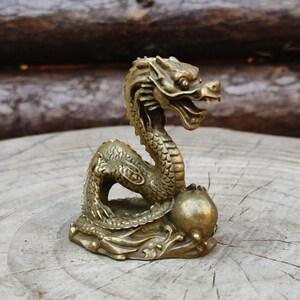 May include: A detailed brass dragon figurine with an open mouth and intricate scales. The dragon is posed on a base with a round object, set against a natural wood background. The sculpture is approximately 4 inches tall.