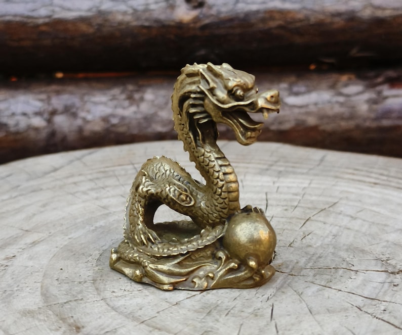 May include: A detailed brass dragon figurine with an open mouth and intricate scales. The dragon is posed on a base with a round object, set against a natural wood background. The sculpture is approximately 4 inches tall.