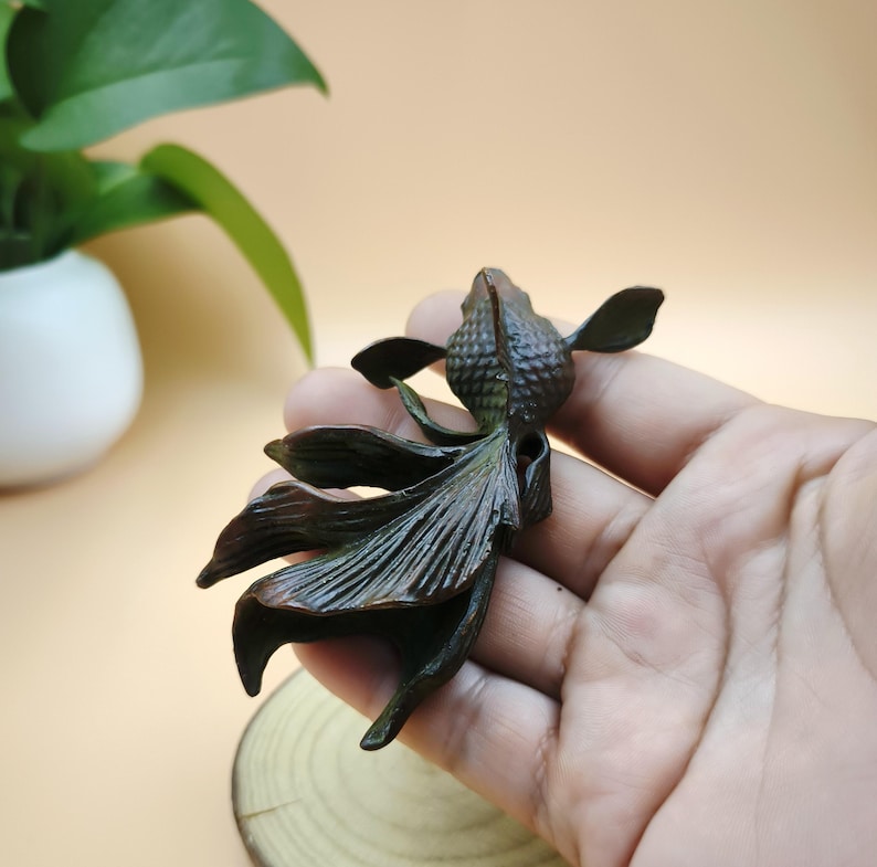 May include: A bronze-colored, detailed sculpture of a goldfish. The fish has flowing fins and a textured body. The sculpture is held in a hand, with a blurred background.