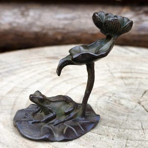 Puede incluir: Una figura de rana de color bronce que descansa sobre una hoja de nenúfar, con una flor encima. La rana está detallada con piel texturizada, y la hoja de nenúfar y la flor tienen vetas intrincadas. La escultura está sobre un fondo de madera natural.