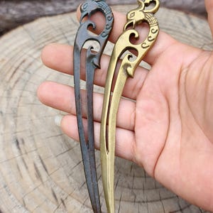 Puede incluir: Dos horquillas decorativas para el cabello. Una es gris oscuro, la otra dorada. Ambas tienen un diseño ornamentado en la parte superior con una figura de pájaro. Las horquillas se sostienen en una mano sobre un fondo de madera borroso.