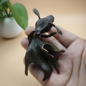 May include: A bronze-colored goldfish figurine with detailed fins and tail. The sculpture has a textured surface and a small, square marking on its back. The figurine is held in a hand, showcasing its size and intricate design.