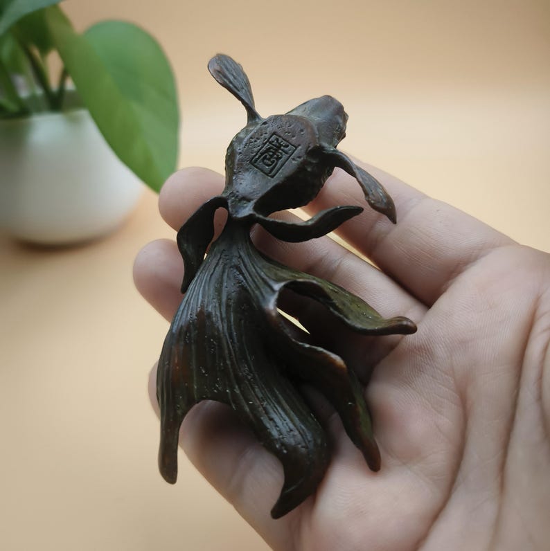 May include: A bronze-colored goldfish figurine with detailed fins and tail. The sculpture has a textured surface and a small, square marking on its back. The figurine is held in a hand, showcasing its size and intricate design.
