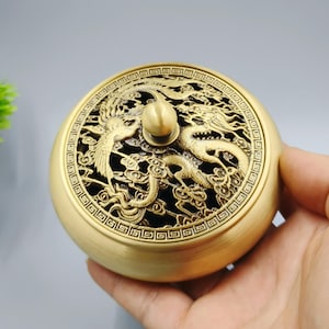 May include: A round, gold-colored incense burner with intricate dragon and phoenix designs on the lid. The lid features a pierced pattern, and a small knob sits atop. The burner is held in a hand.