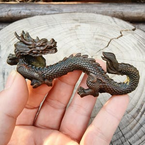 Könnte beinhalten: Detaillierte Bronzedrachenfigur mit aufwendigen Schuppen, Hörnern und einem fließenden Schwanz. Der Drache ist in einer dynamischen, gebogenen Position dargestellt, die seine detaillierten Merkmale zeigt. Die Figur wird in einer Hand gehalten, was ihre Größe und Handwerkskunst hervorhebt.