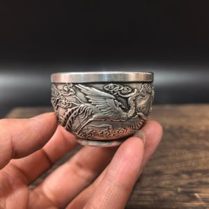 Puede incluir: Una pequeña copa redonda de metal con acabado plateado. La superficie exterior presenta un diseño en relieve detallado que representa una grulla volando rodeada de nubes estilizadas. El borde es liso y sencillo, contrastando con el cuerpo texturizado.