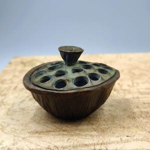 May include: A bronze-colored incense burner with a lid featuring multiple circular openings. The lid is topped with a small, cone-shaped handle. The base of the burner has a textured, ribbed design. The overall aesthetic is antique and decorative.