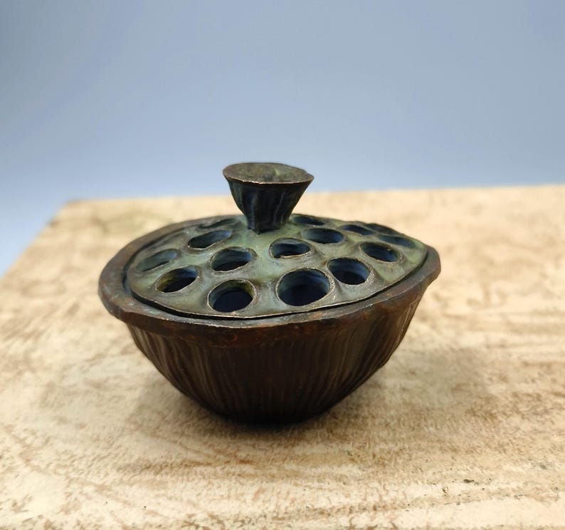 May include: A bronze-colored incense burner with a lid featuring multiple circular openings. The lid is topped with a small, cone-shaped handle. The base of the burner has a textured, ribbed design. The overall aesthetic is antique and decorative.
