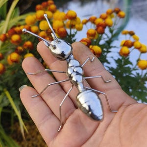 Könnte beinhalten: Eine glänzende, silberne Metallameisenskulptur mit detaillierten Körpersegmenten und dünnen Drahtbeinen und Fühlern. Die Skulptur wird in einer Hand gehalten, mit einem verschwommenen Hintergrund aus gelben und orangen Blumen.