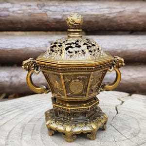 May include: An ornate bronze incense burner with intricate detailing. The lid has a decorative finial and openwork design. The body has handles with dragon head accents and is set on a detailed base. A beautiful piece for home decor.