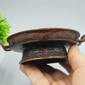 May include: A detailed, antique bronze-colored bowl with ornate, carved patterns. The bowl has a wide, shallow dish with two small handles and a pedestal base. The intricate designs suggest a historical or cultural significance, possibly for decorative or ceremonial use.