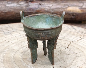 May include: A small, bronze-colored, three-legged vessel with a rounded bowl and two handles. The vessel has intricate decorative patterns around the rim and on the legs. The interior of the bowl has a green patina, suggesting age or exposure to the elements.